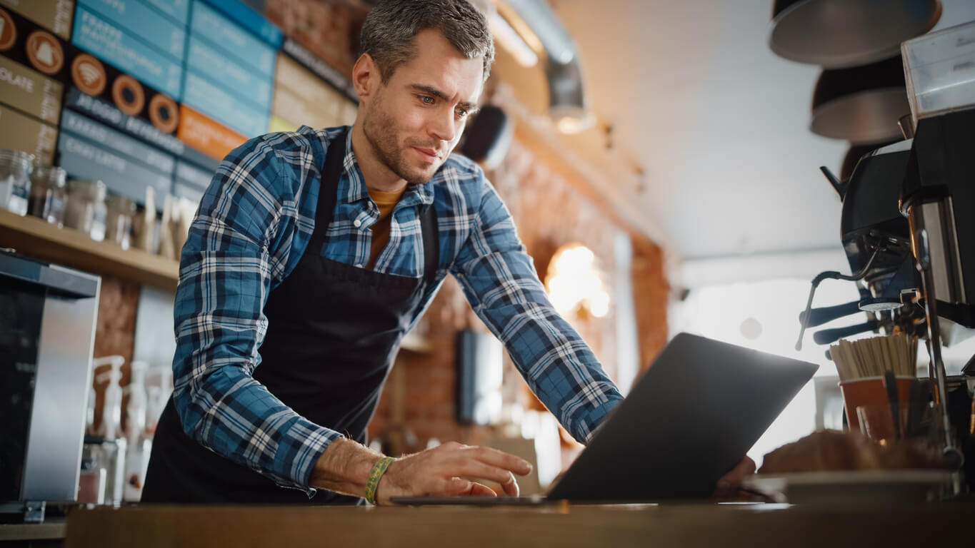 A man wearing a blue plaid shirt and an apron leaning over a workbench next to a coffee machine working on a laptop.