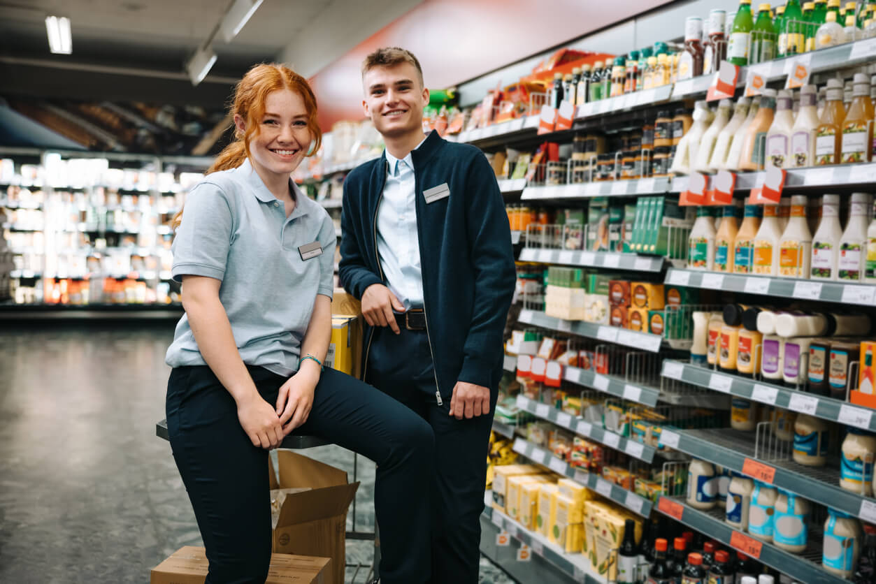 A young man and woman in a grocery store aisle leaning against boxes wearing blue uniforms and name tags and smiling at the camera.