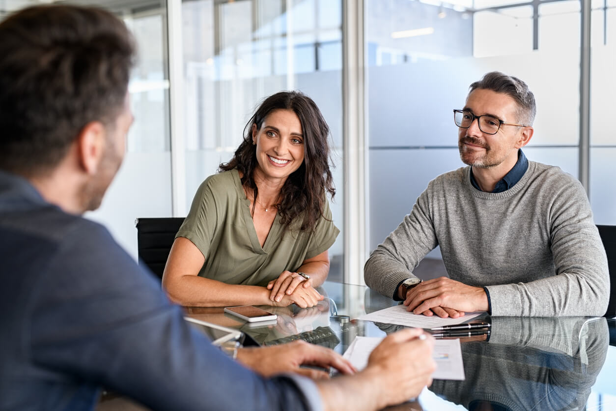 A man and woman sitting in front of paperwork at a glass meeting room table smiling and listening to another man.