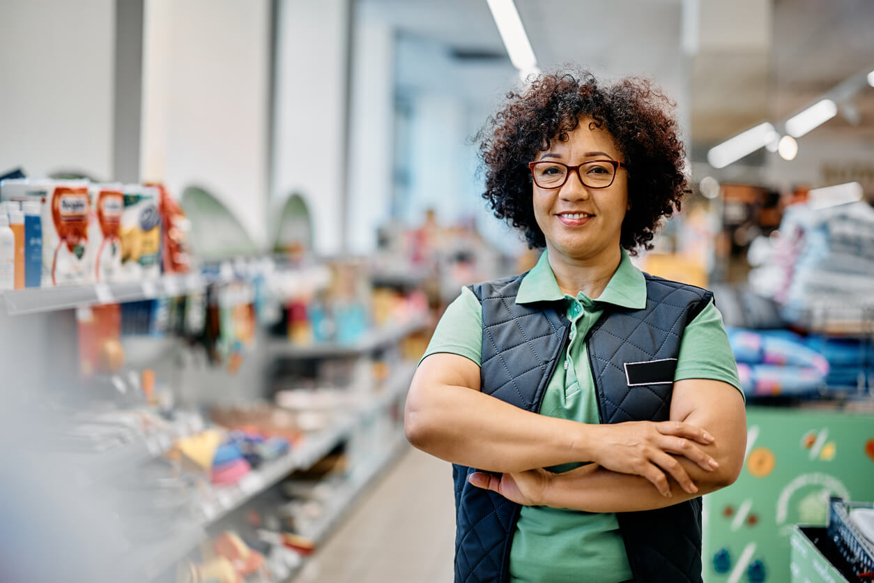 A woman in a grocery store aisle wearing glasses and a uniform of a green polo shirt and a black sleeveless vest crossing her arms and smiling at the camera.