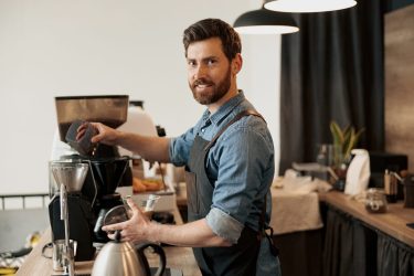 A man wearing an apron pouring coffee beans into a grinder at a cafe smiling.