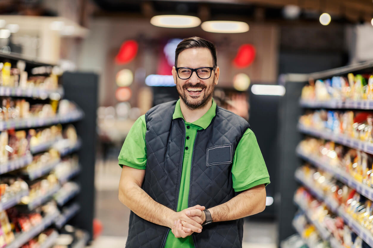 A man standing in a grocery store aisle wearing glasses and a uniform of a bright green polo shirt and a black sleeveless vest smiling widely.