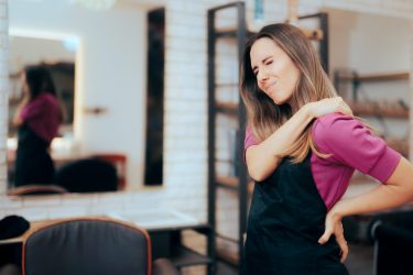 A woman in an apron is closing her eyes and grimacing, holding one hand on her lower back while the other reaches across to rub her shoulder.