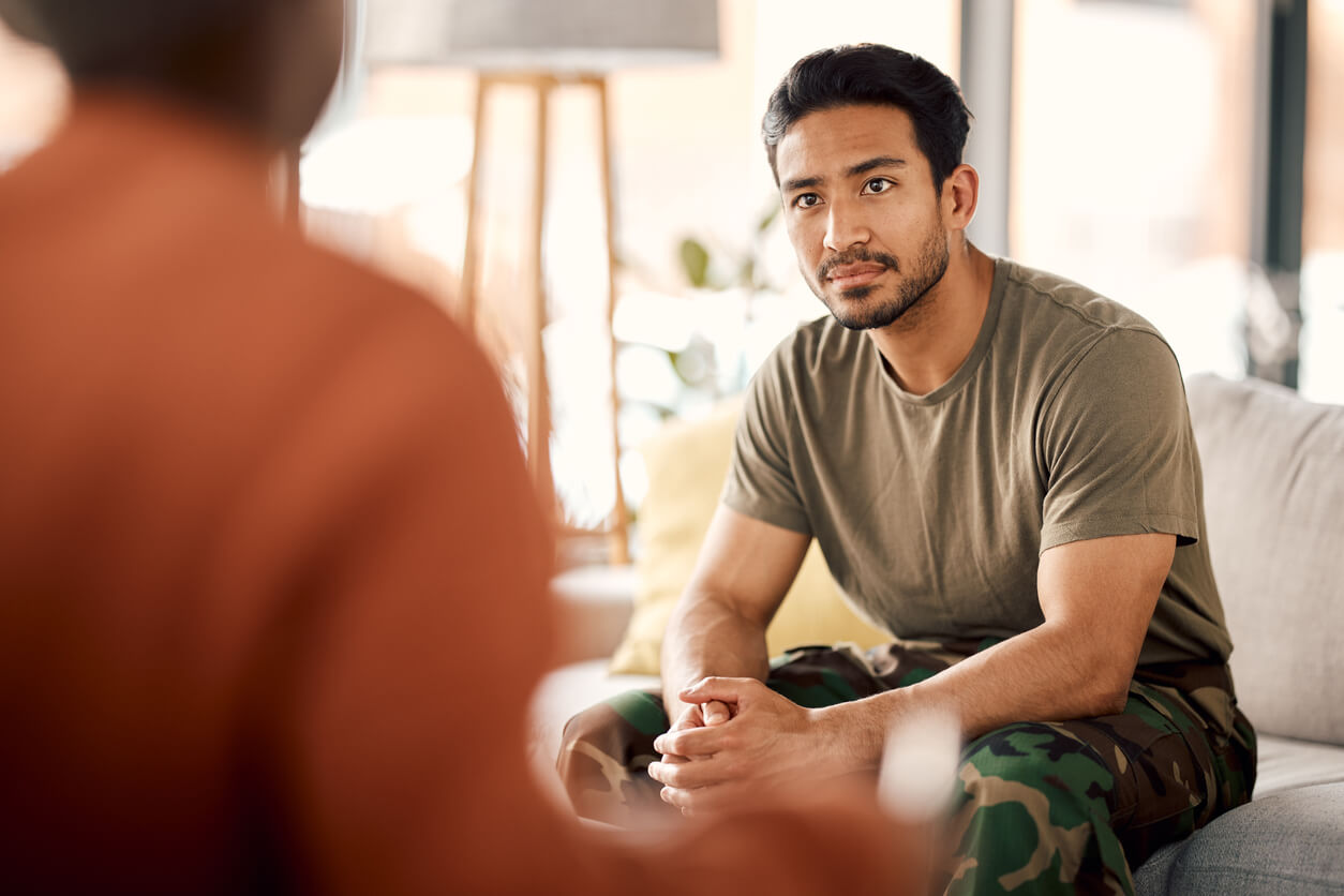 A man wearing a khaki t-shirt and camouflage pants sitting on a sofa with his hands clasped on his lap listening to someone speaking.