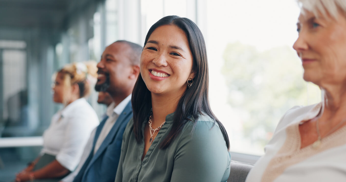 A woman dressed in corporate clothing seated in a row of people smiling and looking at the camera.