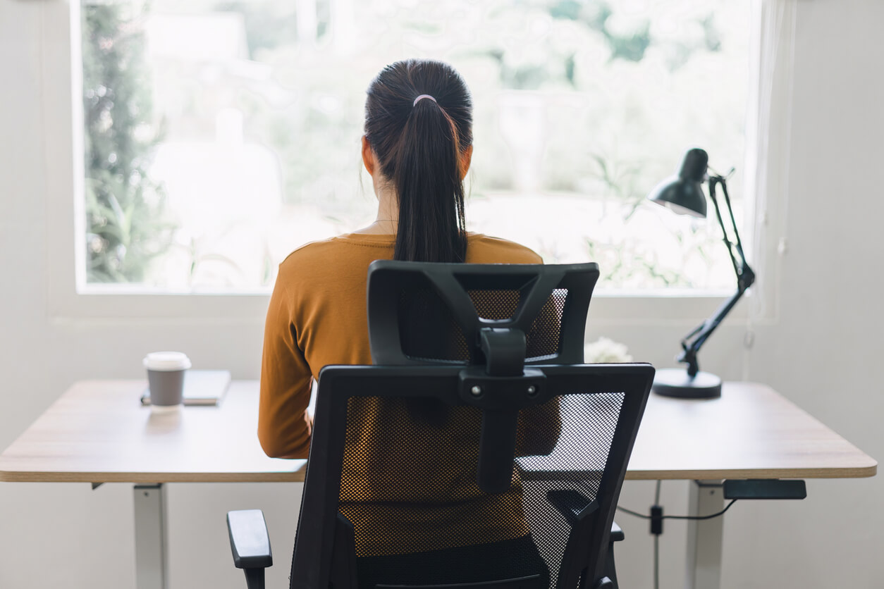 A woman with her back to the camera sitting in an ergonomic chair at a desk by a window.