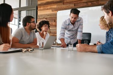 A man leaning over a meeting table problem-solving with a team who are seated around him.