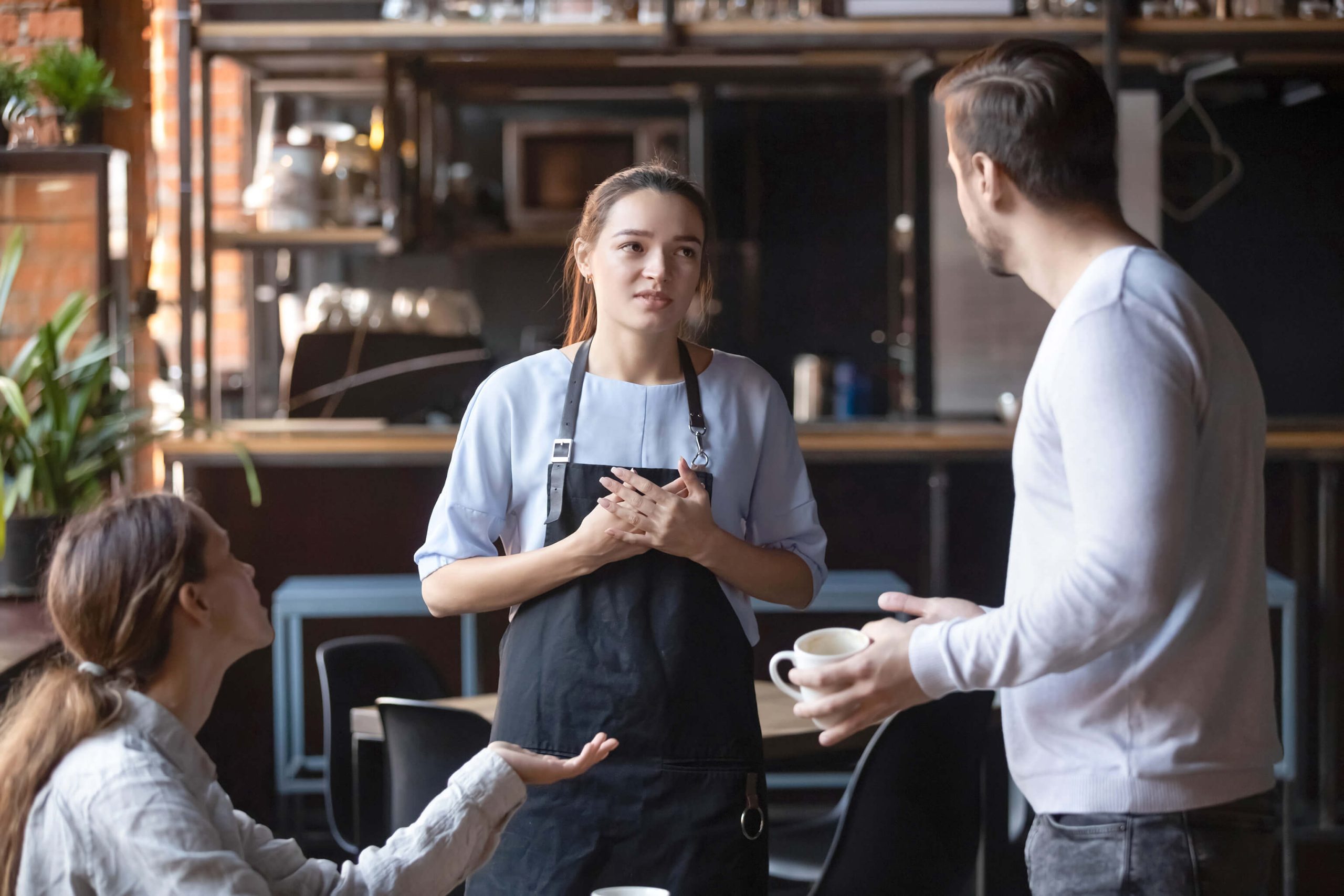 A woman in an apron looking concerned while talking to a couple at a cafe who are gesturing and appearing angry.